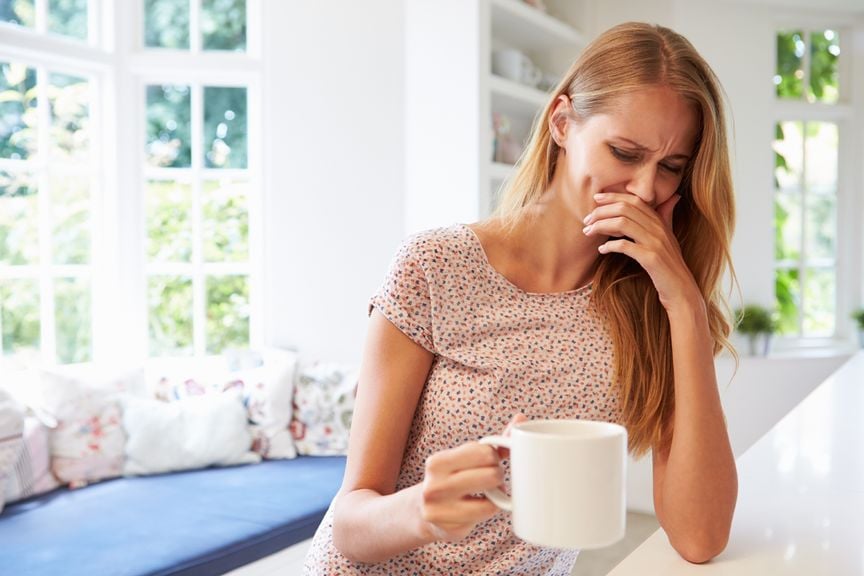 Frau mit einem Becher in der Hand, eine Hand vor dem Mund, verzieht das Gesicht 