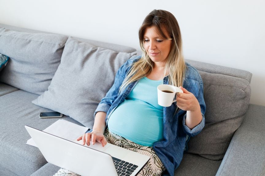 Schwangere mit Laptop sitzt auf dem Sofa und trinkt Kaffee