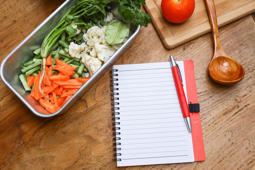 Geschnittenes Gemüse in Lunchbox, daneben ein Notizblock mit Kugelschreiber und eine Holzkelle