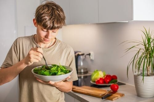 Männlicher Teenager steht in der Küche und isst Salat aus einem Teller