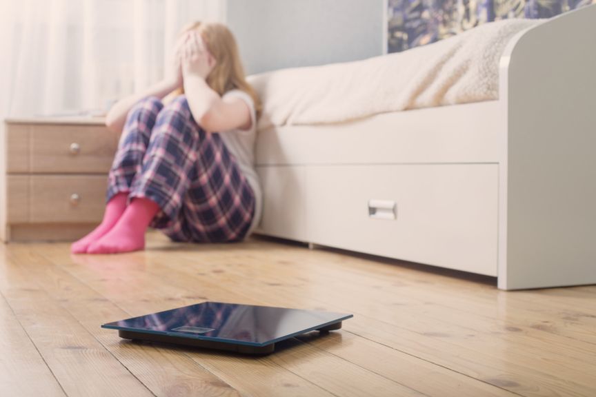 Trauriger weiblicher Teenager sitzt auf dem Fussboden mit einer Personenwaage vor sich