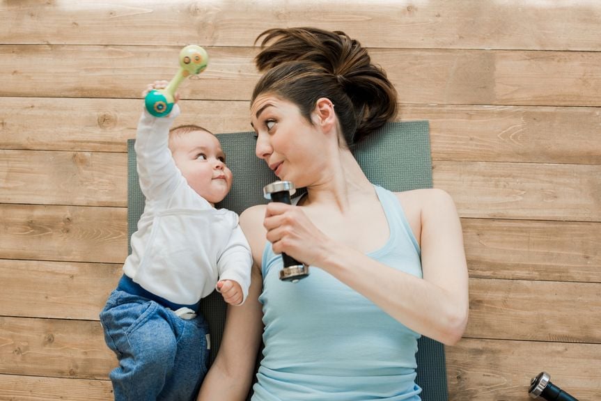 Mutter liegt mit Baby auf einer Fitnessmatte, beide mit einer Hantel