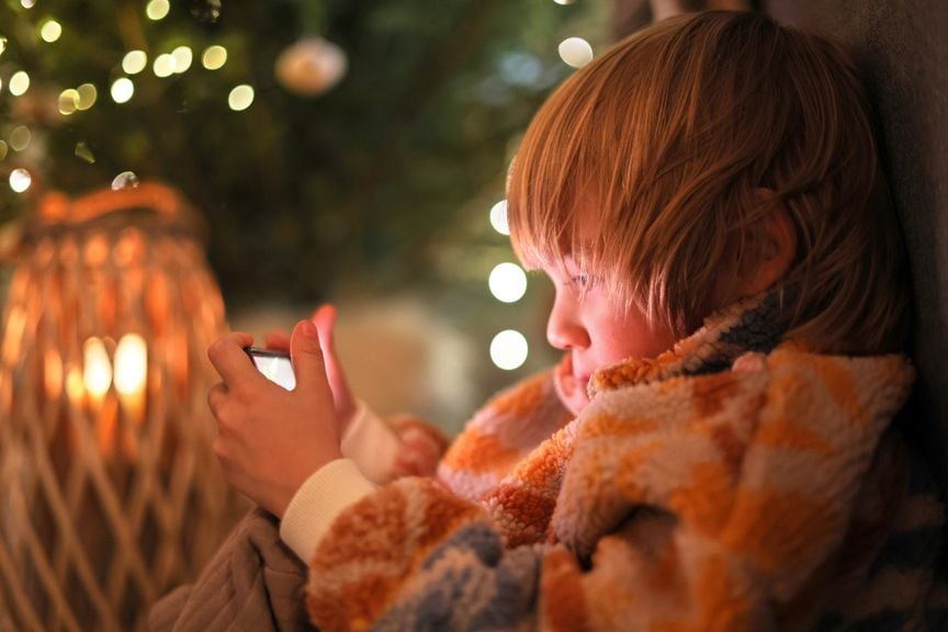 Kind sitzt in weihnachtlich dekoriertem Raum und schaut aufs Smartphone