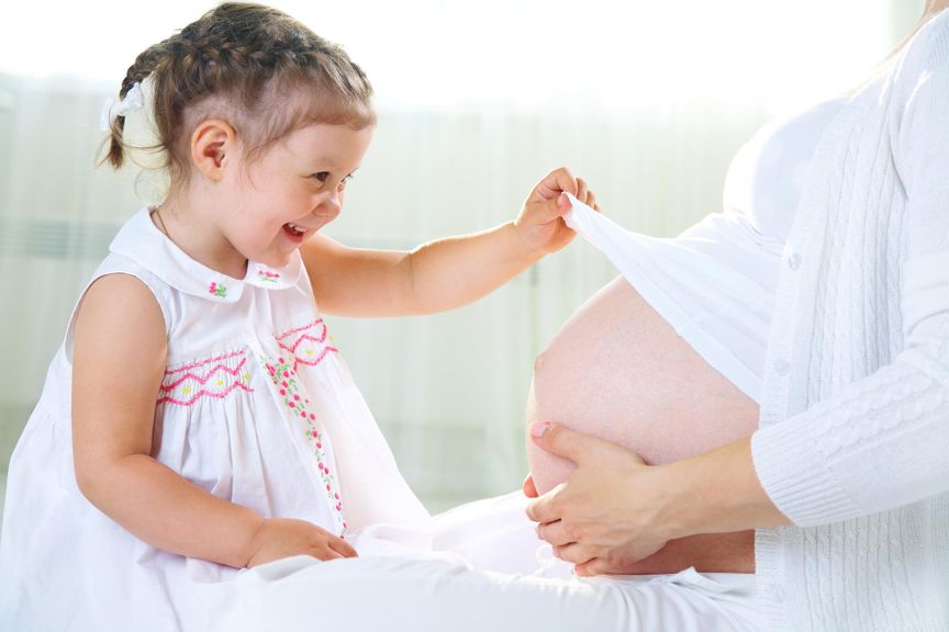 Mädchen hebt der Mutter das T-Shirt und stahlt den Babybauch an