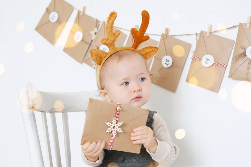 Baby mit Rentier-Haarreif hält ein Adventskalender-Couvert in den Händen