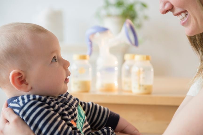 Baby schaut aufmerksam die Mutter an, im Hintergrund abgepumpte Muttermilch in Flaschen