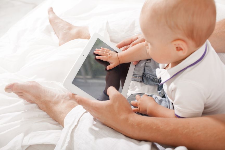 Baby sitzt mit Vater auf dem Bett und schaut auf ein Tablet