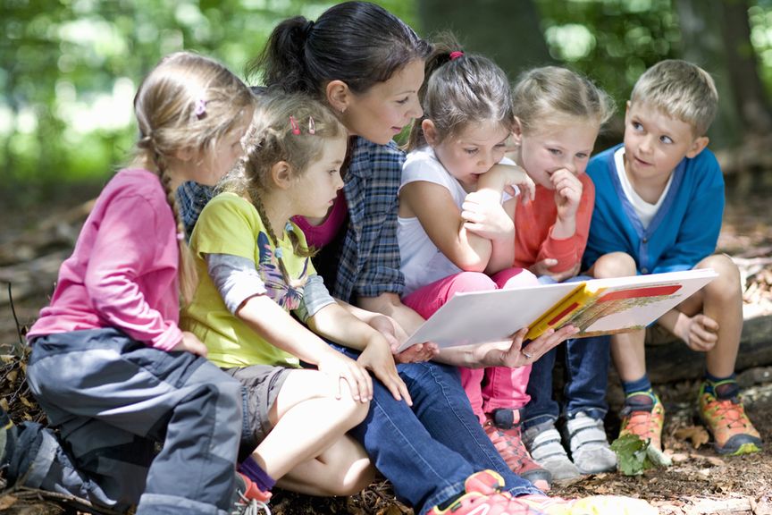 Kindergärtnerin liest einer Kindergruppe im Wald aus einem Buch vor