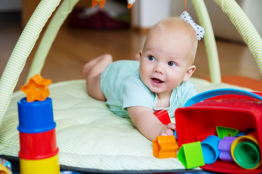 Baby liegt auf dem Bauch unter einem Spielbogen und greift nach dem Spielzeug