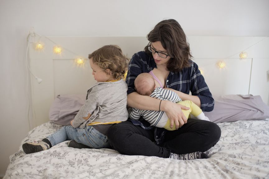 Mutter stillt auf dem Bett, das Geschwisterchen lehnt sich an sie