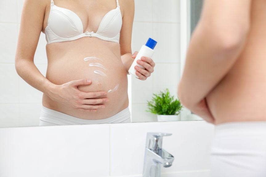 Schwangere cremt sich vor dem Spiegel den Bauch ein
