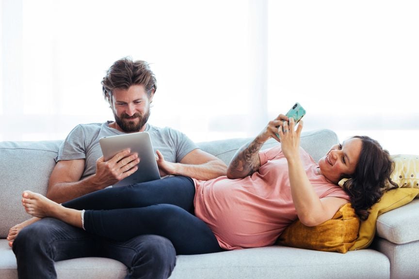 Werdende Eltern auf dem Sofa mit Handy und Tablet