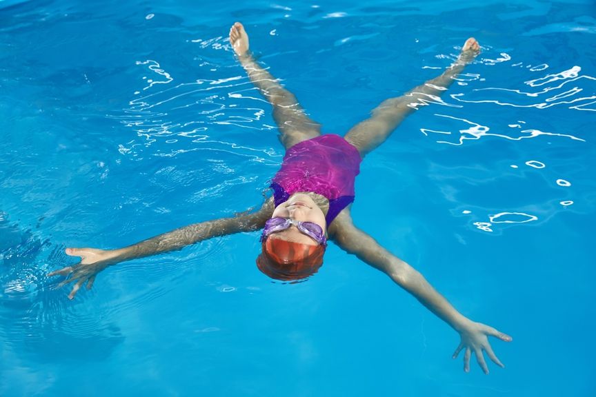 Kind mit Schwimmbrille liegt mit ausgestreckten Armen und Beinen im Wasser und macht den "Seestern"