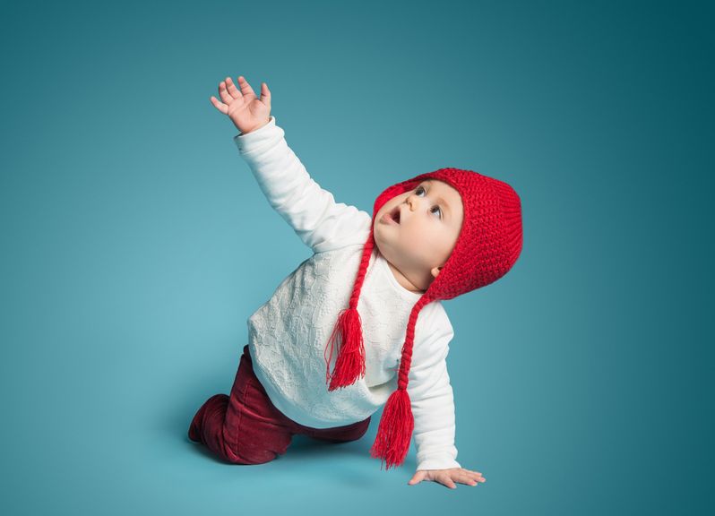 Baby mit Mütze krabbelt am Boden und streckt einen Arm hoch