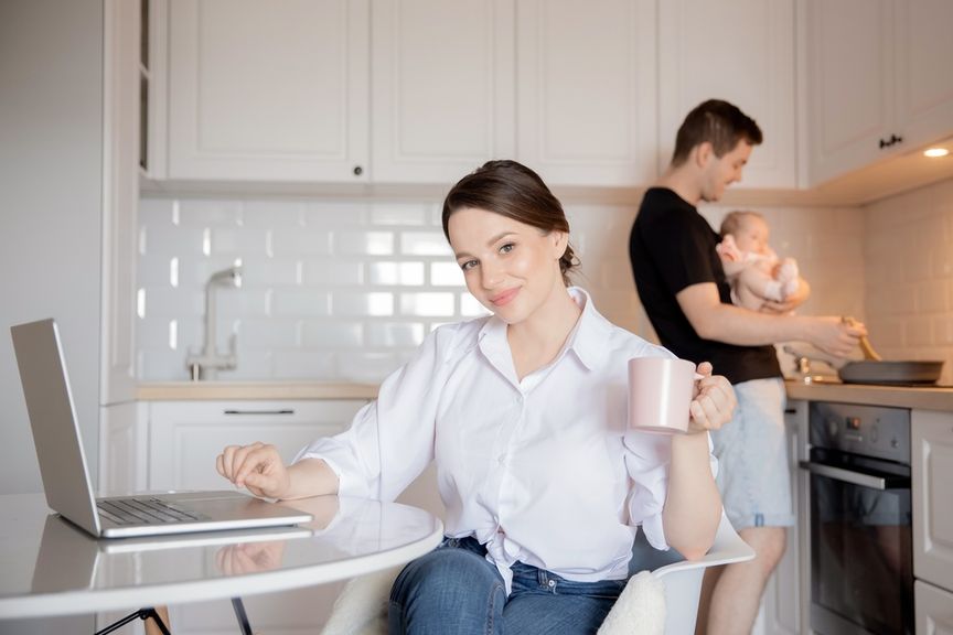 Frau arbeitet in der Küche am Laptop, Mann kocht mit dem Baby auf dem Arm