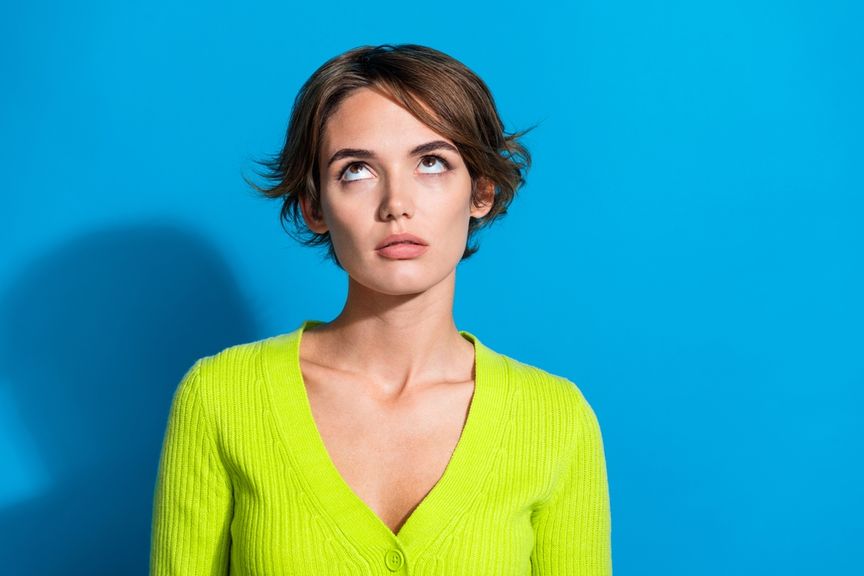 Eine kurzhaarige Frau im neongrünen Pullover steht vor einen türkisfarbenen Hintergrund und verdreht die Augen