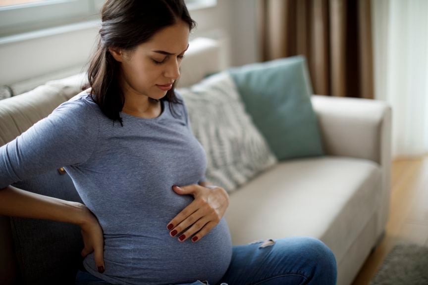 Schwangere sitzt auf dem Sofa und hält sich Bauch und Rücken