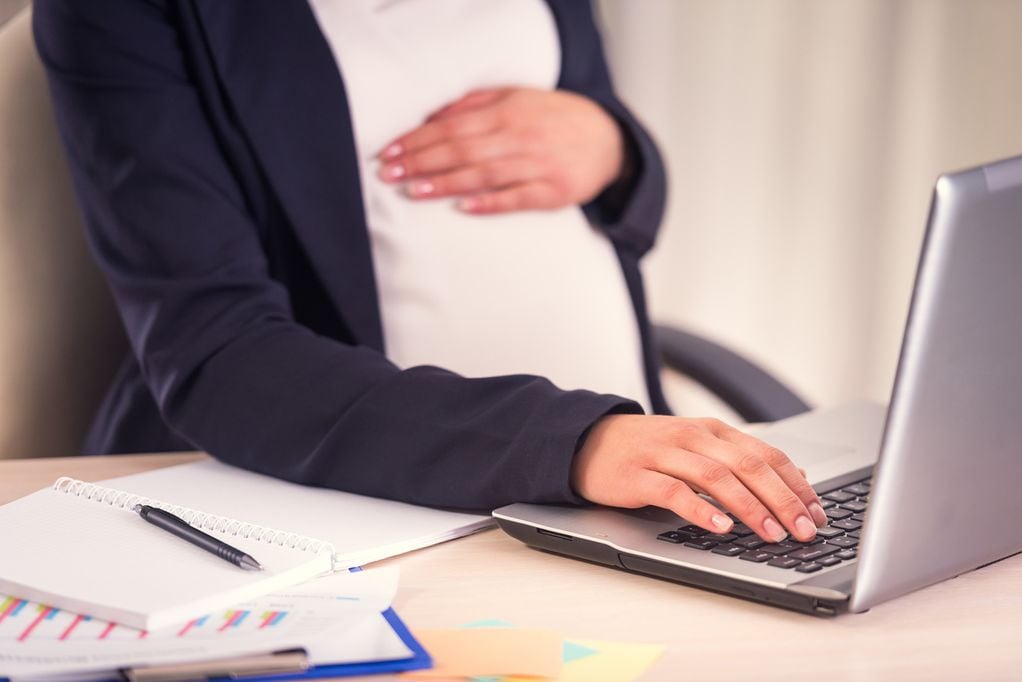 Schwangere am Schreibtisch, die Hand auf dem Bauch, mit Laptop