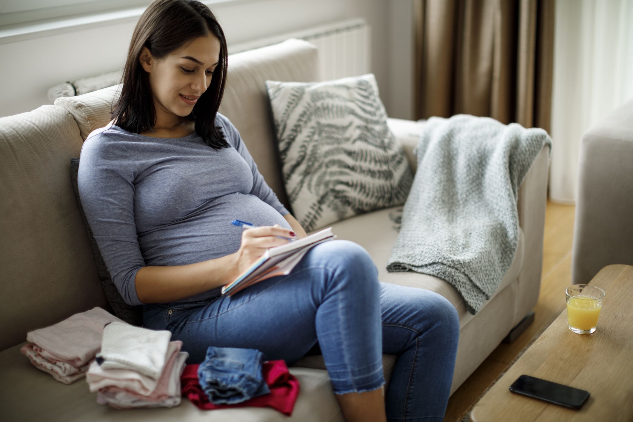 Schwangere mit Notizizettel und Babykleidung auf dem Sofa
