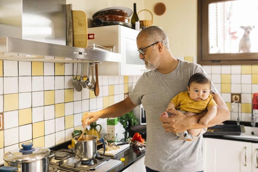 Älterer Vater mit einem Baby auf dem Arm steht am Herd und kocht