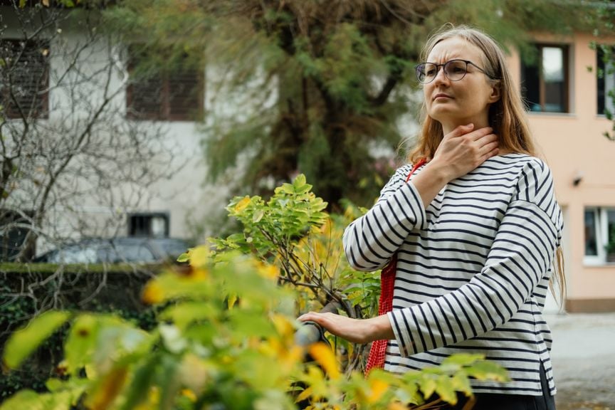 Frau mit Einkaufswagen steht draussen, greift sich an den Hals und schaut verunsichert. 