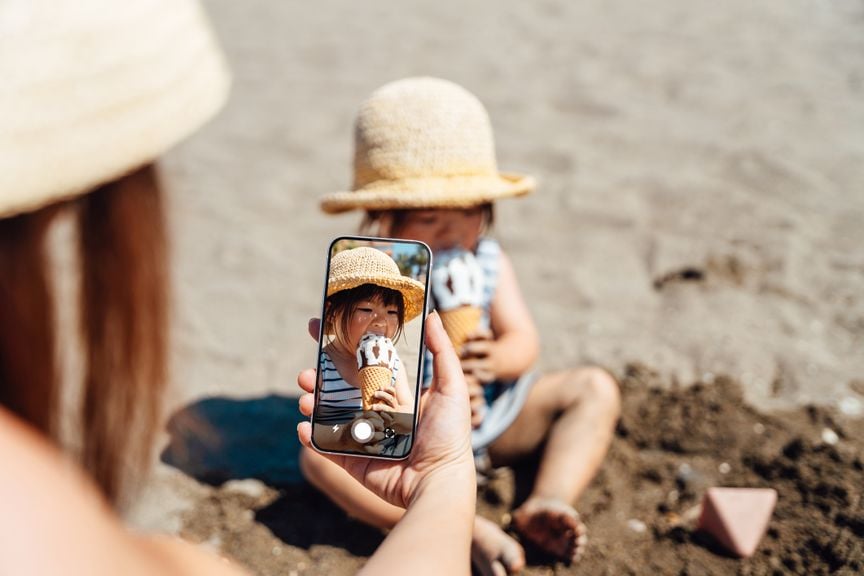 Frau fotografiert mit dem Handy ein Kleinkind, das am Strand ein Cornet isst