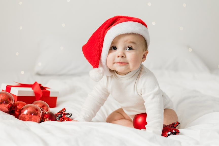 Baby in einem weissen Body mit einer Nikolausmütze sitzt im Bett mit Geschenk und Weihnachtsdekorationen