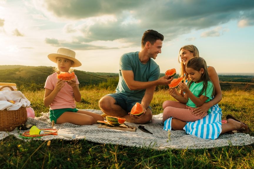 Familie mit zwei Kindern sitzt in idyllischer Umgebung auf einer Picknickdecke und isst Wassermelone