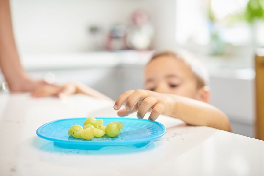 Kind greift nach Weintrauben, die auf einem Plastikteller liegen