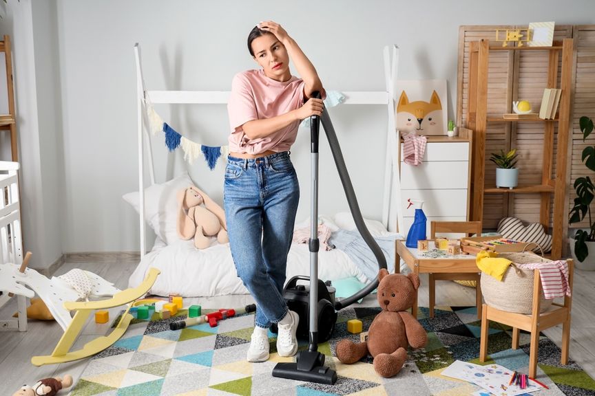 Müde aussehende Frau steht mit Staubsauger in einem unaufgeräumten Kinderzimmer