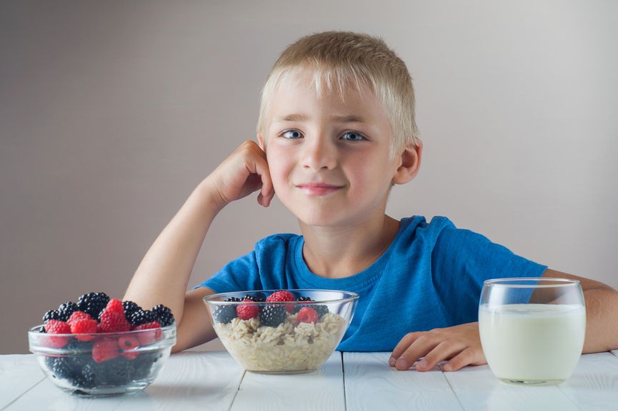 Kind sitzt am Tisch, vor sich ein Glas Milch, eine Schale mit Himbeeren und Brombeeren und ein Müesli mit Beeren