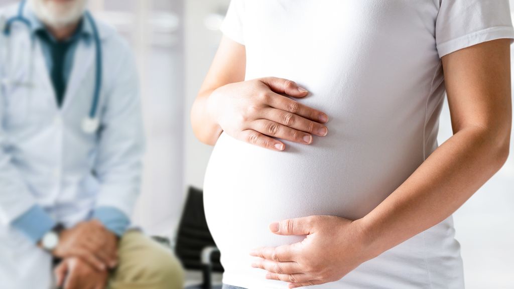 Schwangere hat die Hände vor dem Bauch, im Hintergrund sitzt ein Arzt