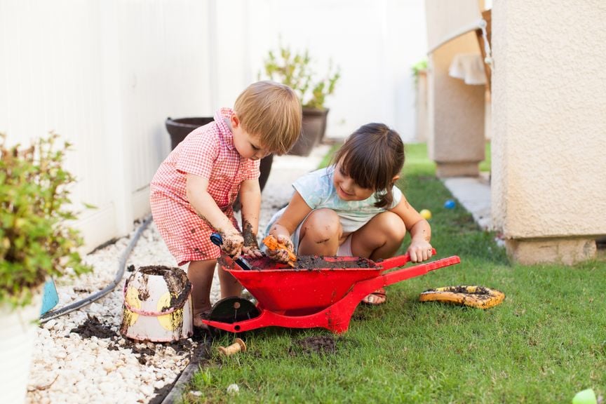 Zwei Kinder schaufeln im Garten Schlamm aus einer Kinder-Schubkarre