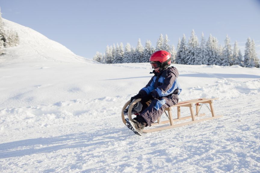 Kind mit Helm und Skibrille schlittelt auf einem Holzschlitten in verschneiter Landschaft