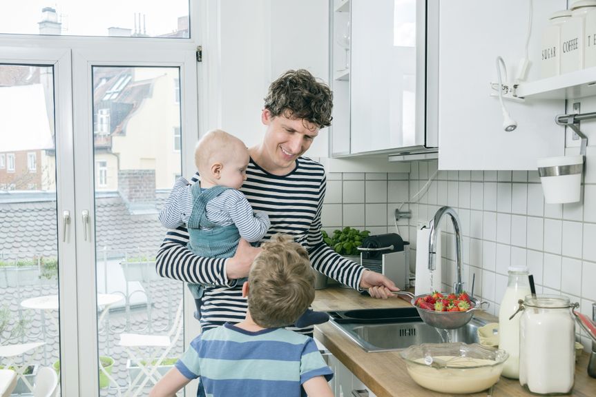 Vater hält ein Baby auf dem Arm, spült Erdbeeren ab und spricht mit seinem Sohn