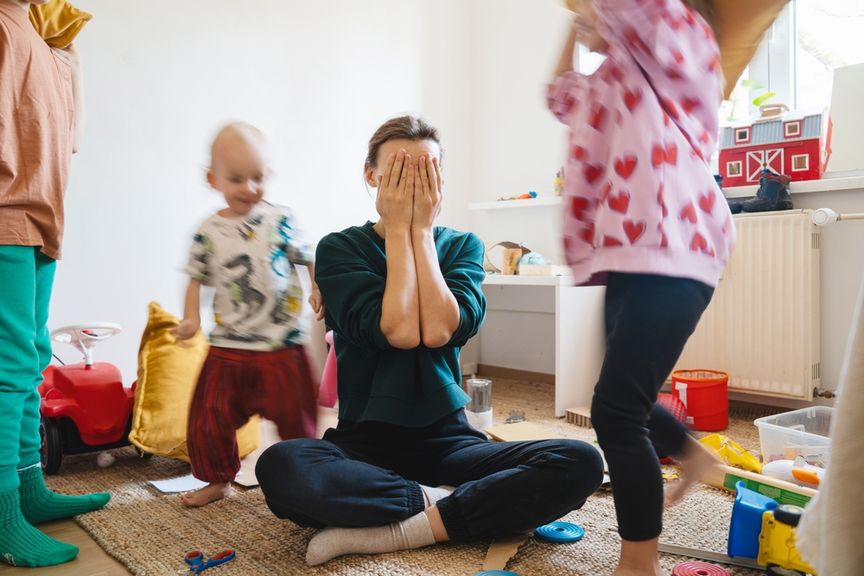 Überforderte Mutter sitzt im Kinderzimmer und Kinder rennen um sie herum