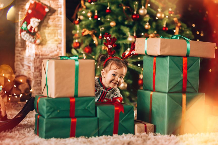 Kleinkind schaut lächelnd hinter aufgetürmten Weihnachtsgeschenken hervor
