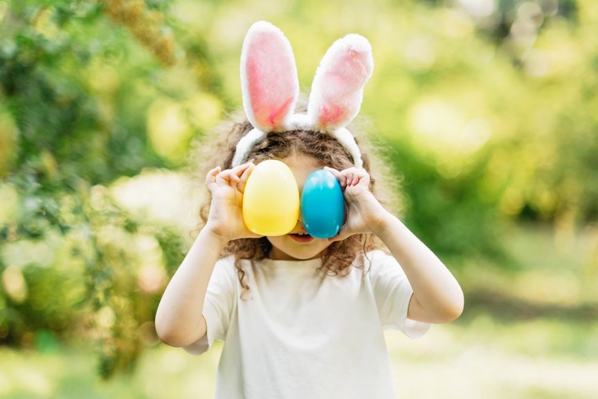 Kind mit Hasenohren-Haarreif hält sich ein gelbes und ein blaues Osterei vors Gesicht