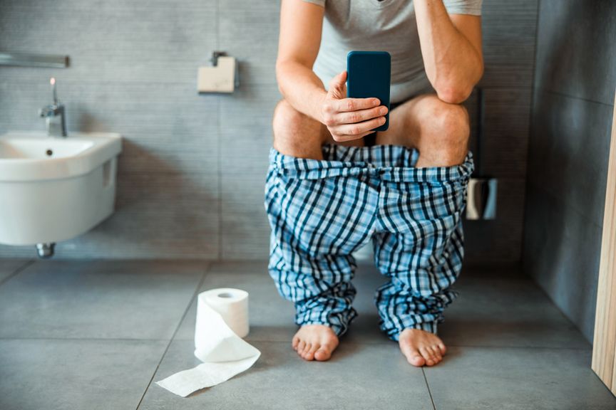 Mann in Pyjamahose sitzt mit dem Handy in der Hand auf der Toilette