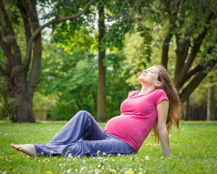 Schwangere Frau sitzt entspannt auf einem Rasen umgeben von Bäumen.