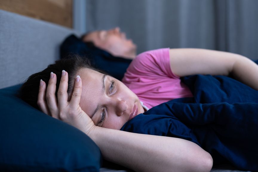 Frau liegt neben schlafendem Mann schlaflos im Bett