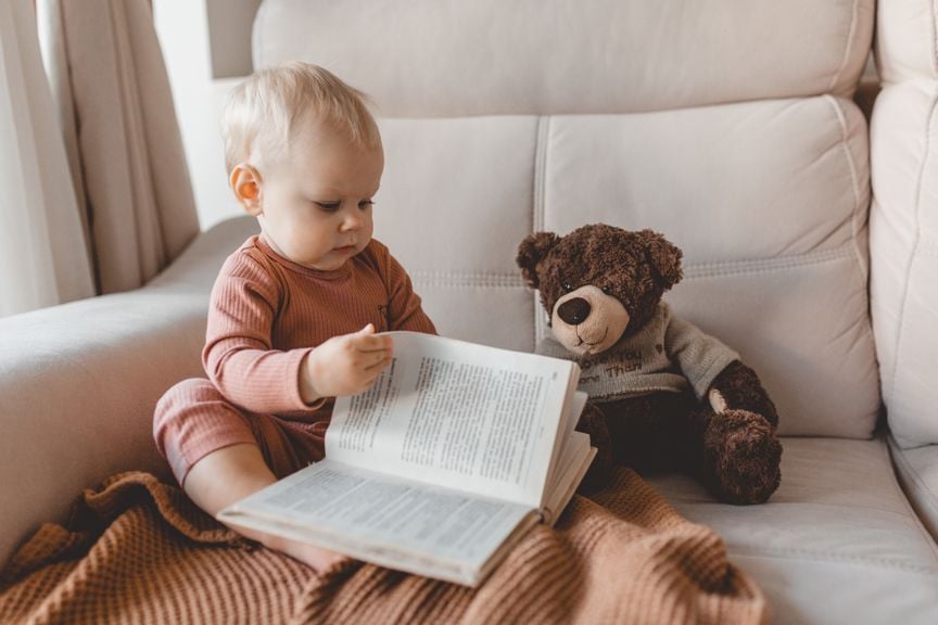 Kleinkind blättert in einem Buch, Teddybär sitzt daneben