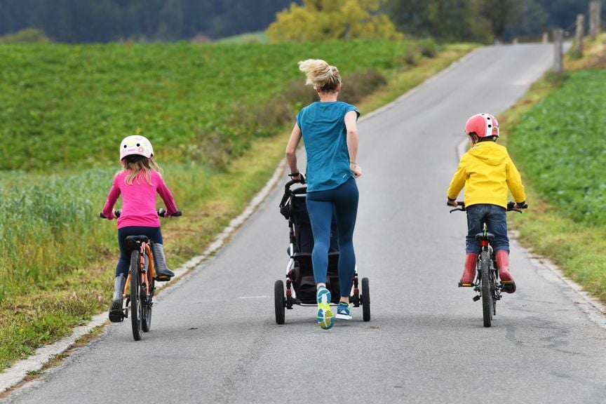 Frau joggt mit Kinderwagen und zwei Kindern auf dem Velo