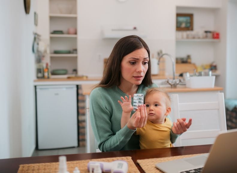 Junge Frau hält Blisterpackung mit Tabletten in der Hand, Baby auf dem Schoss und schaut auf den Laptop