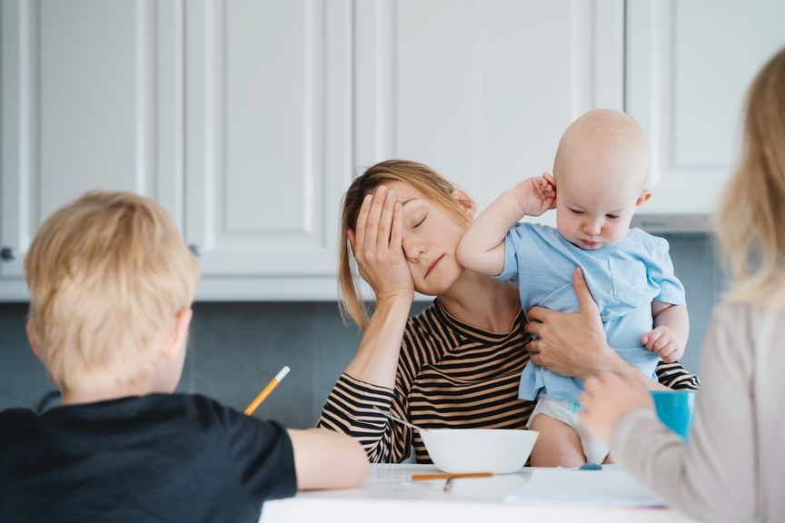 Übermüdete Mutter mit Baby auf dem Arm sitzt am Frühstückstisch, wo ein grösseres Kind seine Hausaufgaben macht