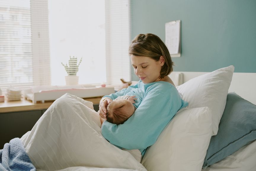 Mutter mit Baby auf dem Arm sitzt auf Kissen gestützt im Bett und stillt