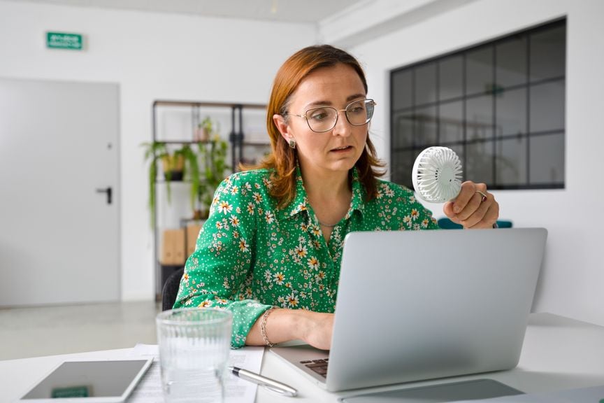 Frau mit Hitzewallungen sitz am Laptop und hält einen elektrischen Mini-Ventilator in der Hand