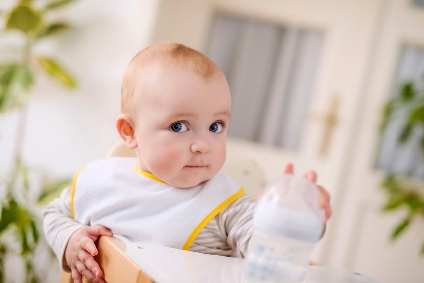Baby sitzt am Tisch, hÃ¤lt eine Wasserflasche und schaut neugierig in die Kamera
