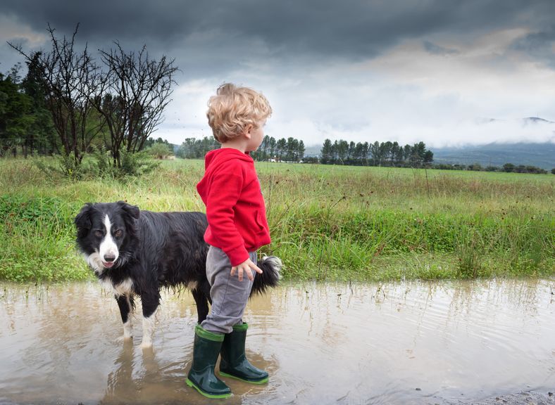 Kind steht mit Hund in einer Wasserpfütze, am Himmel dunkle Gewitterwolken