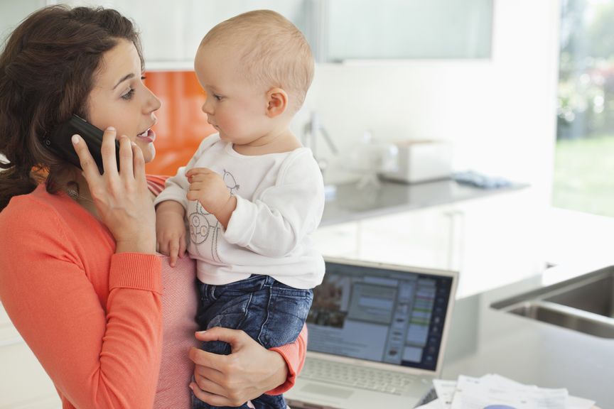 Mutter mit Baby im Arm telefoniert, im Hintergrund der Laptop